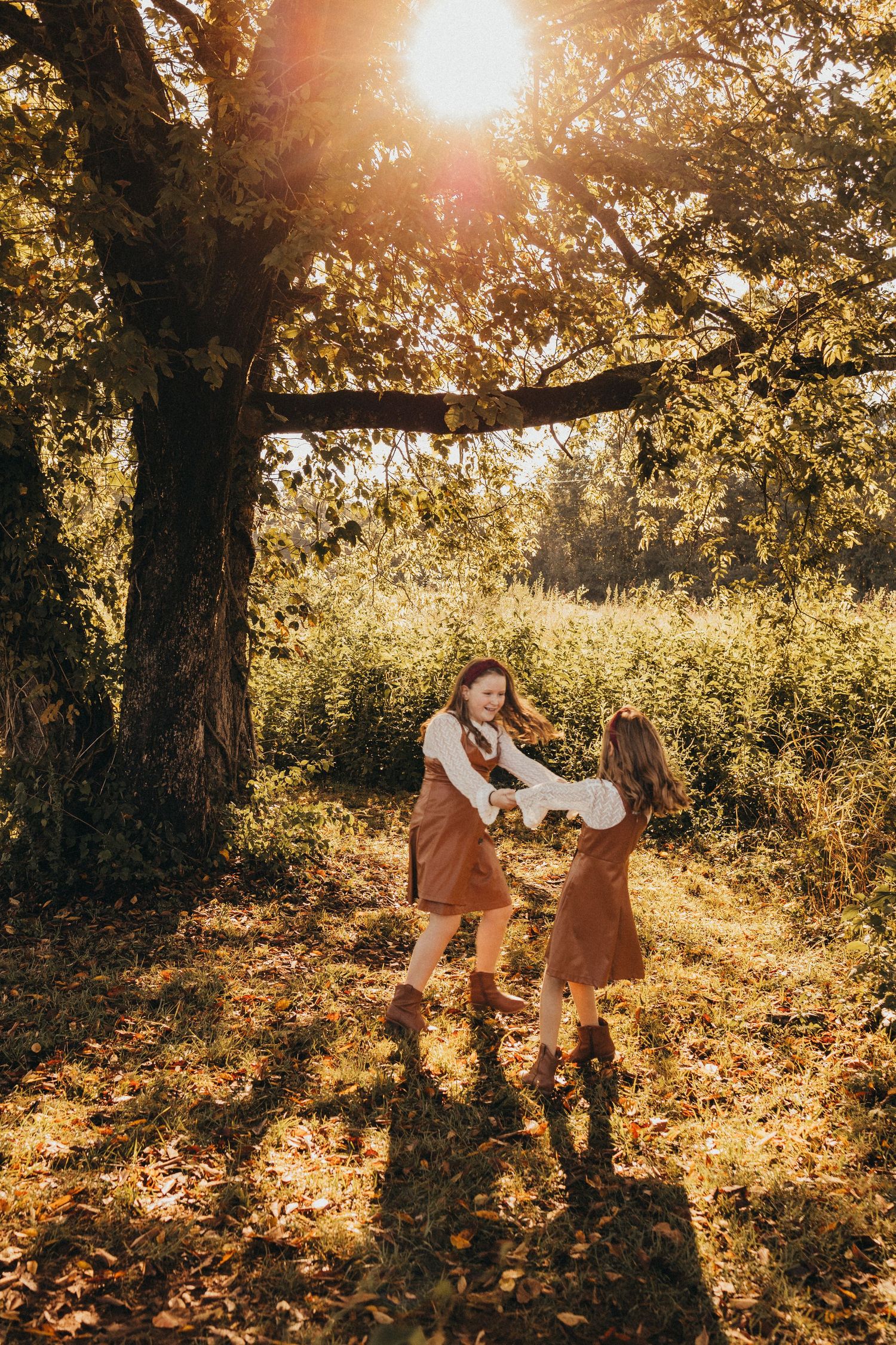 Two girls in matching brown dresses dance playfully together in golden autumn sunlight.