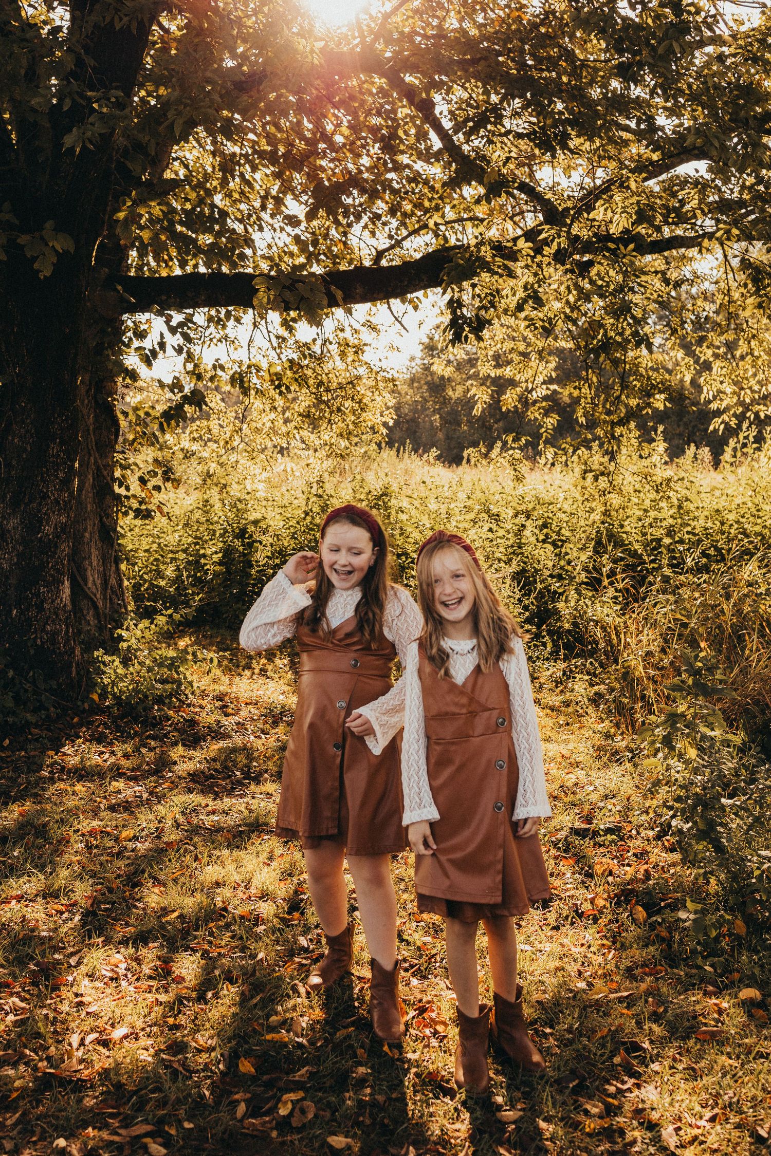 Two cheerful girls in matching brown dresses laugh together under a sunlit autumn tree.
