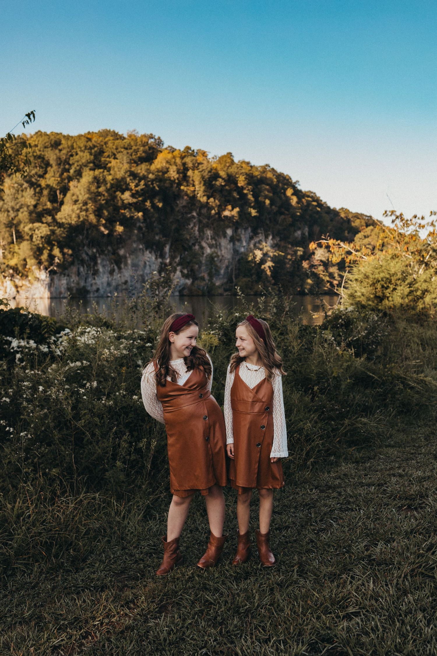 Two girls in matching brown jumper dresses laughing together in a field with a rocky bluff backdrop.
