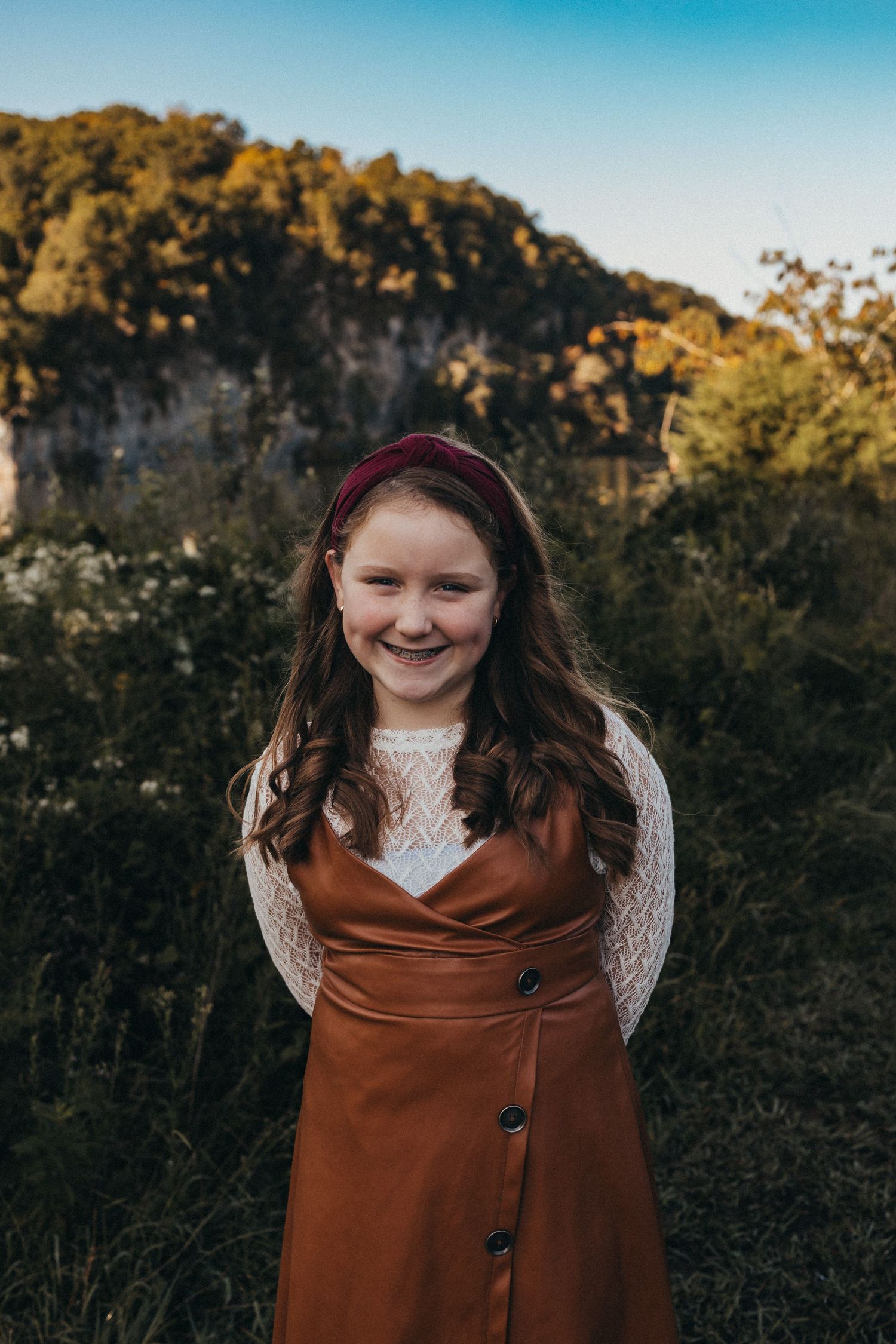 Smiling young girl in a brown dress and burgundy headband standing outdoors with autumn foliage behind her.