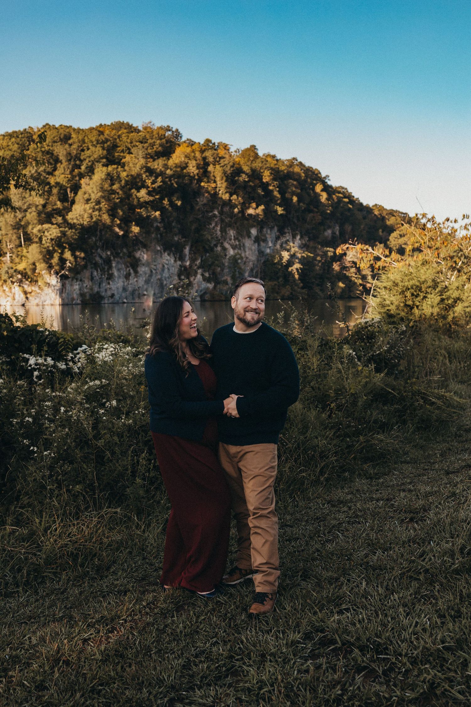 Couple holding hands and smiling at each other outdoors with a rocky bluff and lake in the background.