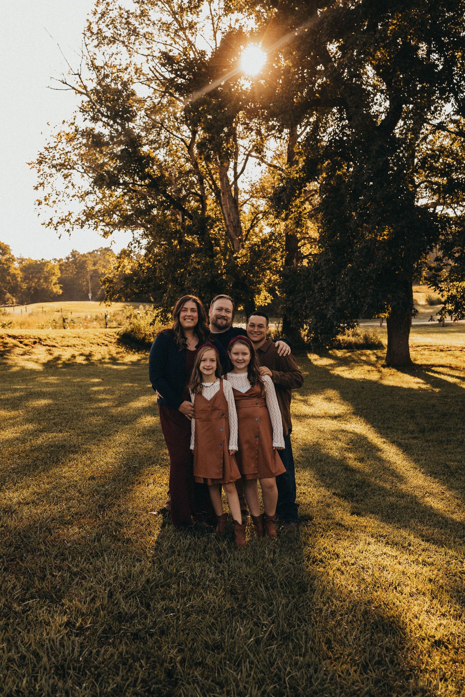Family of five posing together under a large tree with golden sunlight streaming through autumn leaves.
