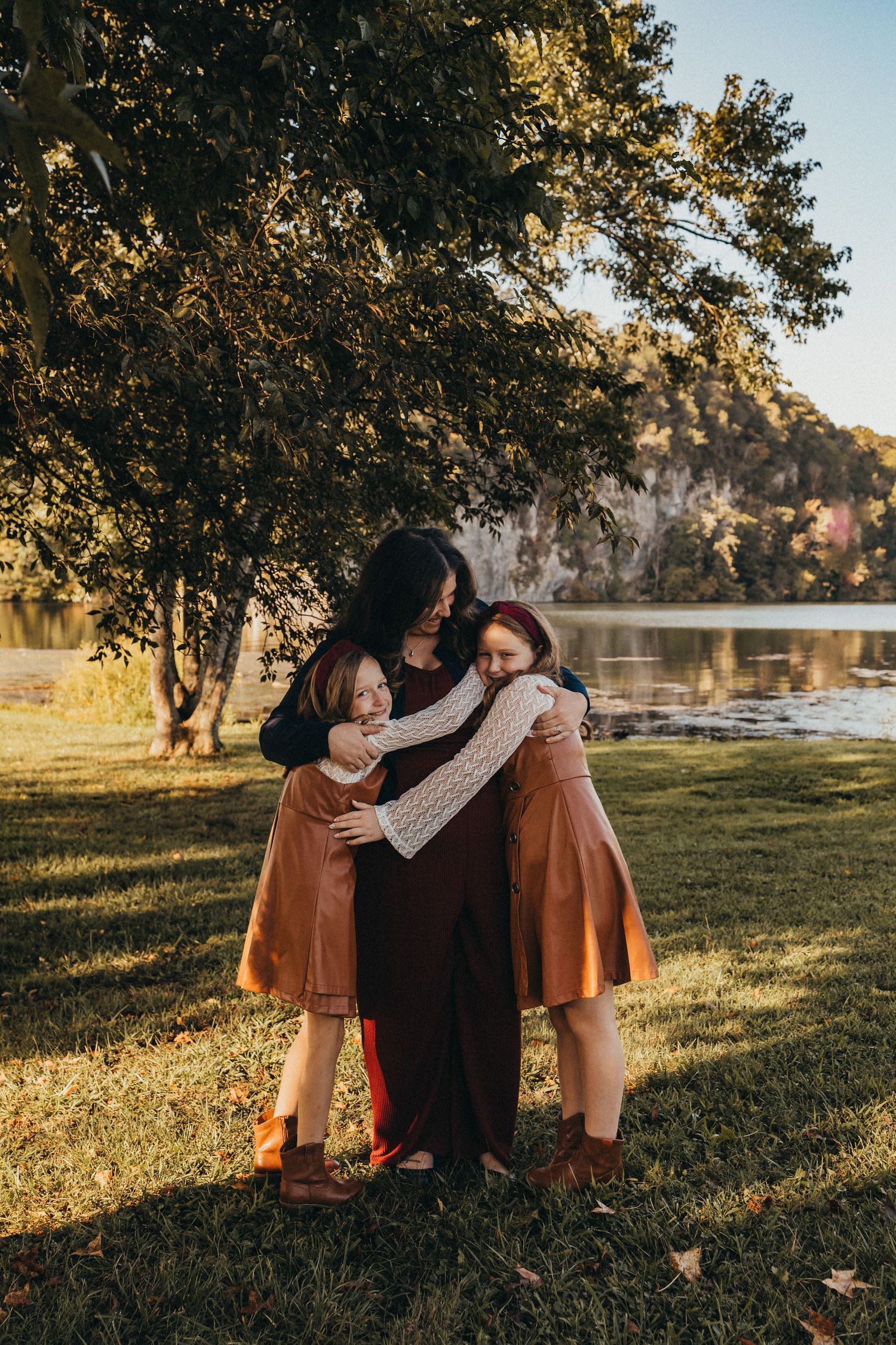 Mother hugging her two daughters under a large tree near a sunlit lake in autumn.