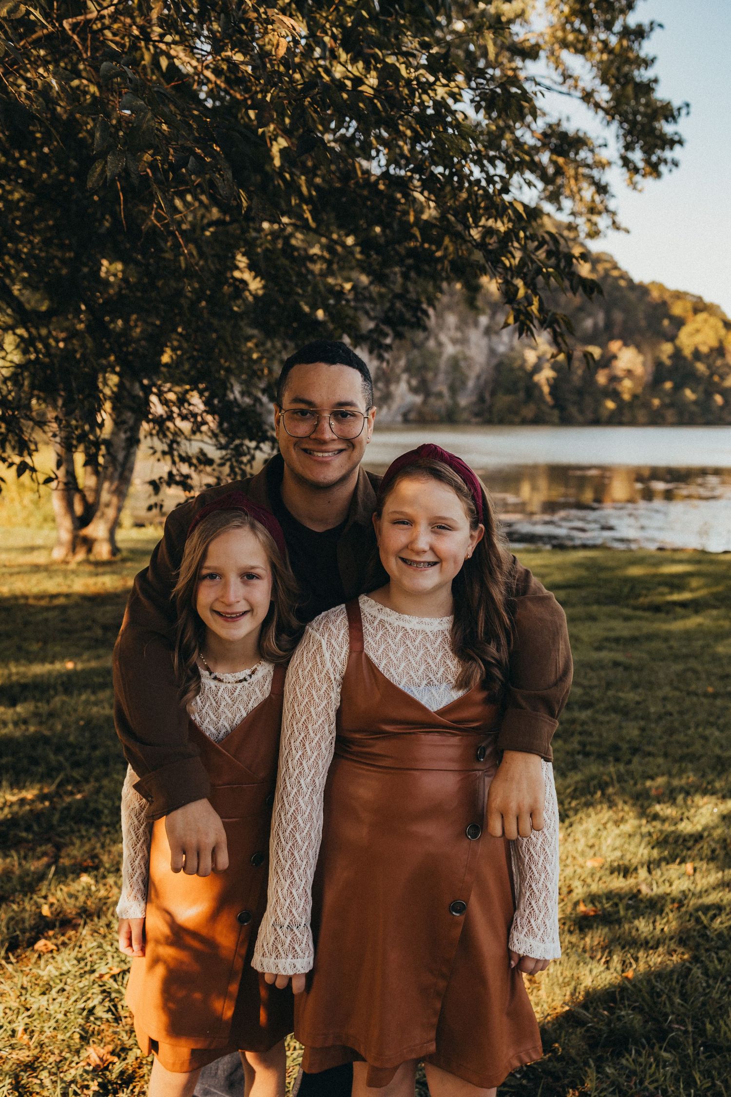 Young man smiling with two girls in matching brown dresses under a tree near a sunlit lake.