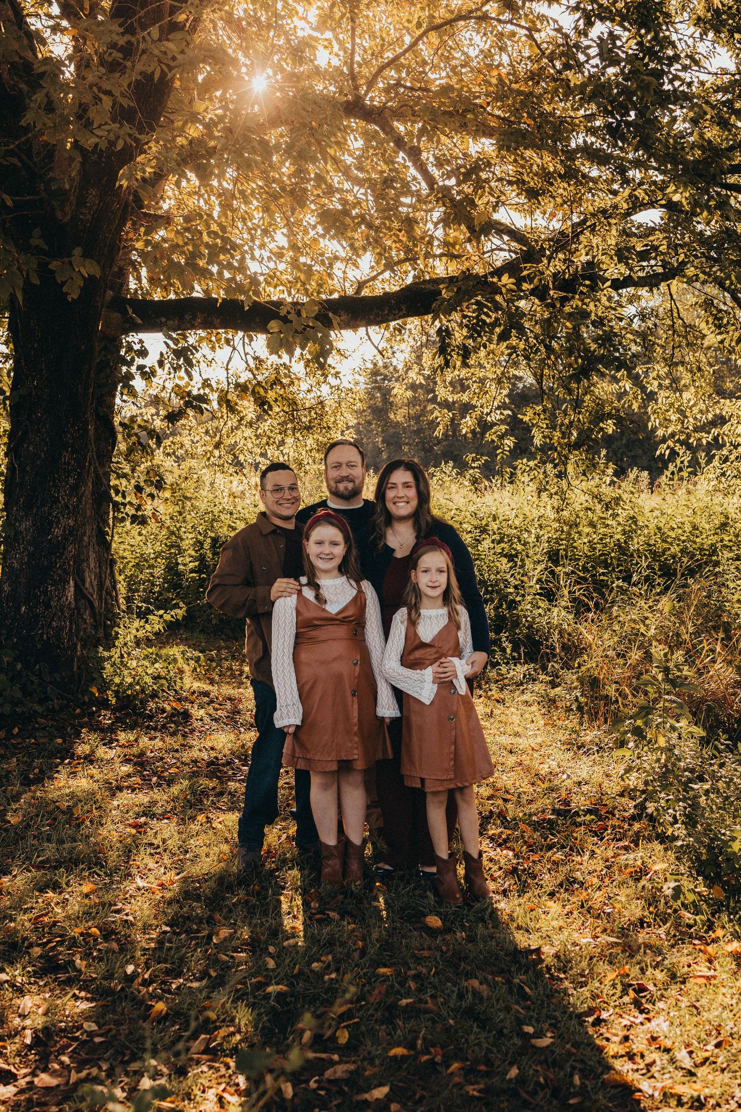 Family of five posing under a golden autumn tree surrounded by lush fall foliage in warm sunlight.