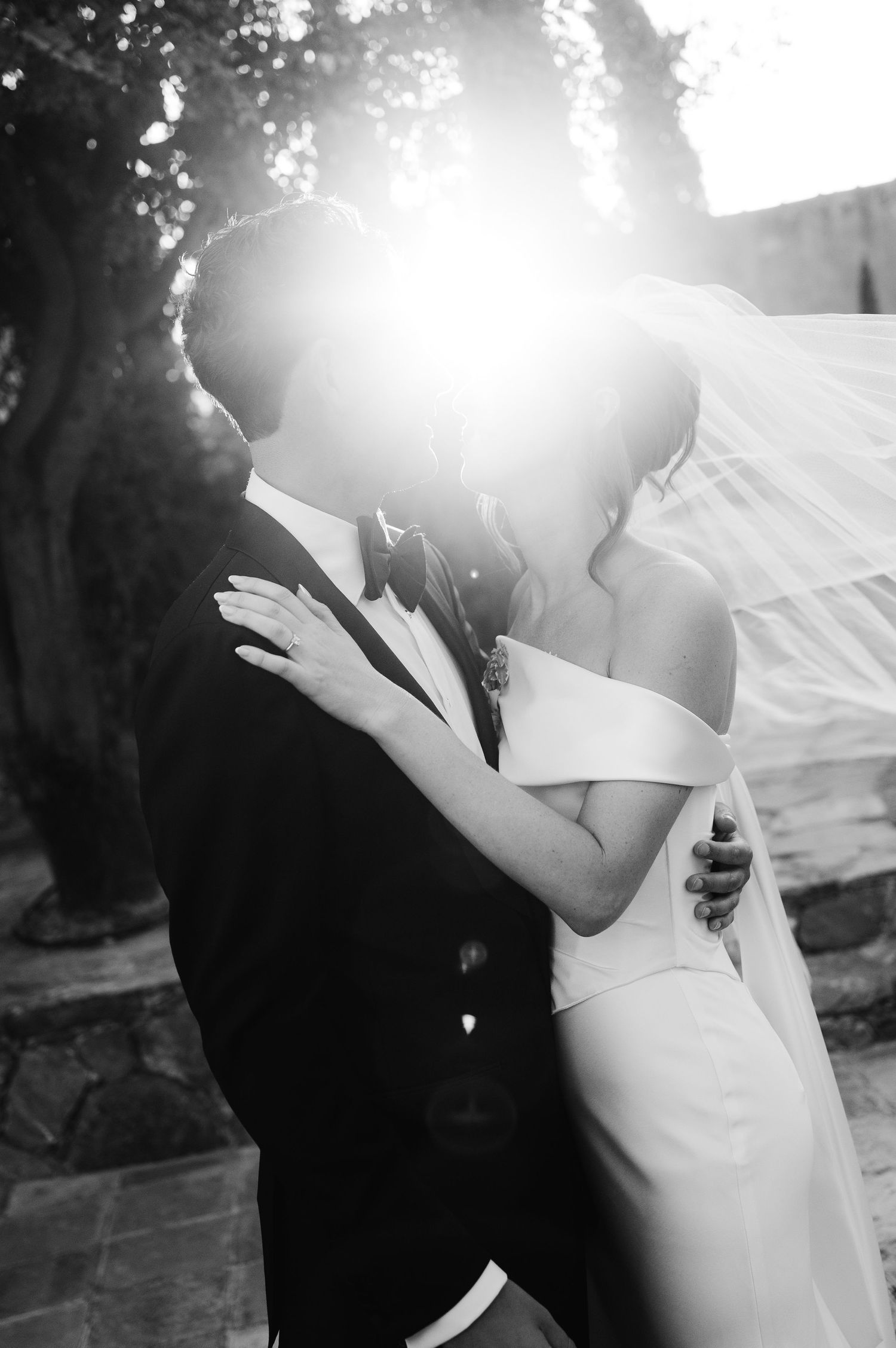 Romantic black and white wedding photo of couple dancing together in sunlit outdoor setting.