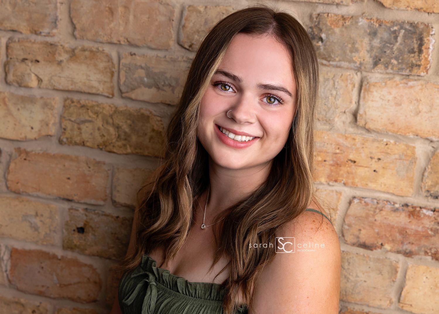 A bright and cheerful portrait of someone with long brown hair smiling in front of a rustic brick wall.