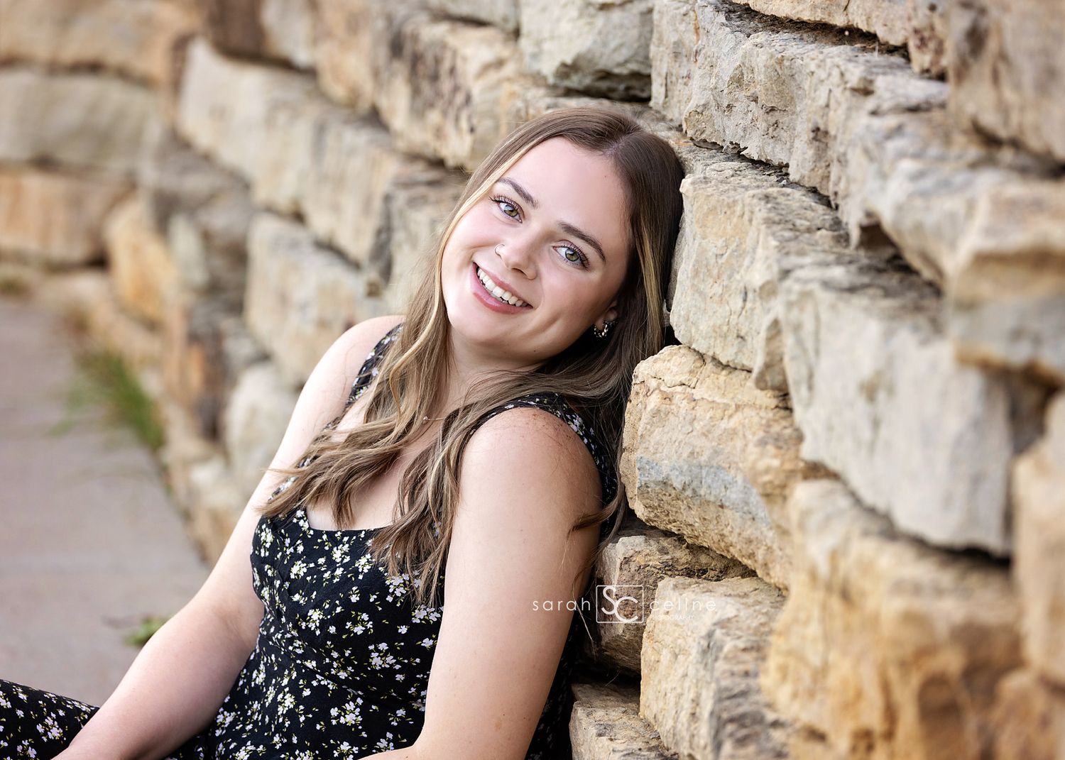 A person in a floral dress poses smiling against a rustic stone wall background.