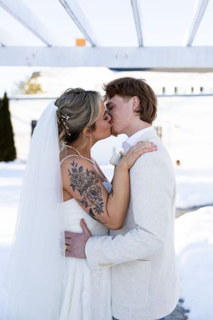 Romantic wedding couple embraces in white attire against bright backdrop, with visible floral tattoo on shoulder.