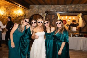 Wedding party in emerald green velvet dresses poses together at rustic indoor venue with stone walls and warm lighting.