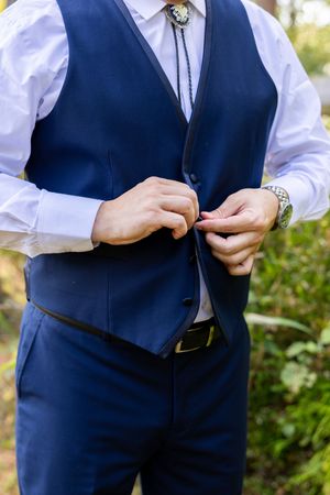 A close-up view of a navy blue vest and white dress shirt with a bolo tie being buttoned.