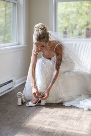 A bride in a lace wedding dress fastening her white heels in a sunlit room.