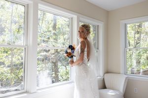 A bride in a white lace gown holding a blue and peach bouquet by large windows.