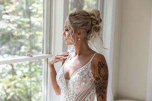 A bride in an elegant white lace dress with intricate details standing by bright windows.