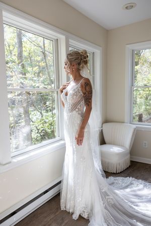 A bride in a form-fitting white lace gown poses by large windows in a bright room.