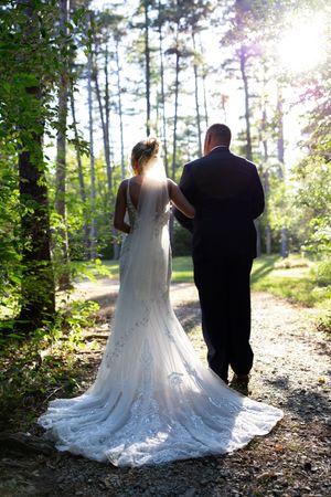 A couple walks hand in hand down a sunlit forest path in wedding attire.