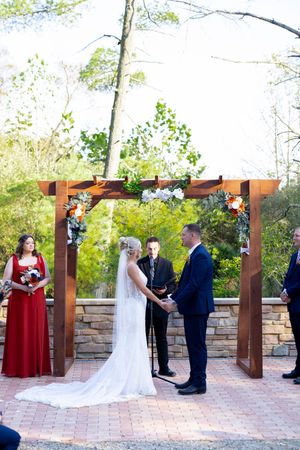 Wedding ceremony under wooden pergola with stone base and decorative floral arrangements.