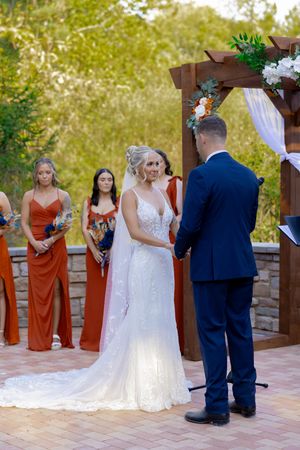 Wedding ceremony with bridesmaids in rust-colored dresses standing beside wooden pergola.