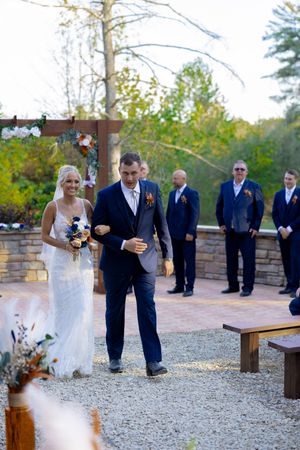 Wedding ceremony processional on gravel path through wooded outdoor venue.