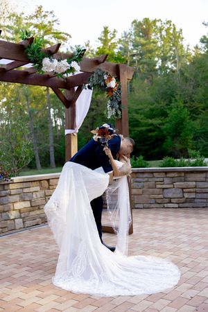 A romantic wedding moment under a decorated wooden pergola with flowing white dress train on stone patio.