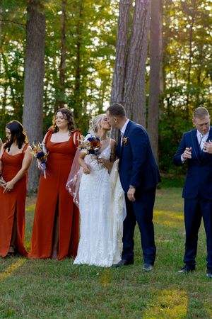 Wedding party in rustic outdoor setting with bridesmaids in rust orange dresses and groomsmen in navy suits.