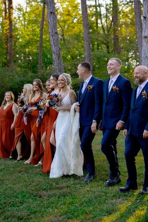 Wedding party poses together on grass with bridesmaids in orange dresses and groomsmen in navy suits.