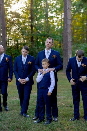 Groomsmen in navy suits stand with young ring bearer in outdoor forest setting.