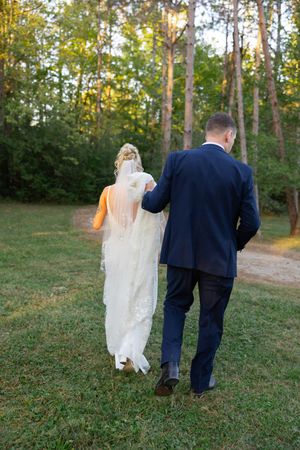 A couple walks together on a grassy path through trees during a wedding photoshoot.