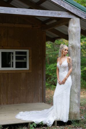 A detailed white lace wedding gown is displayed against a rustic wooden cabin backdrop.