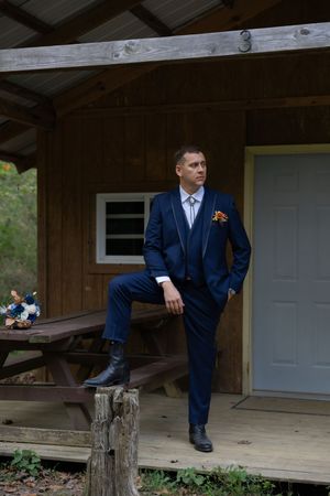 A person in a navy suit sits thoughtfully on a wooden porch during a wedding photoshoot.