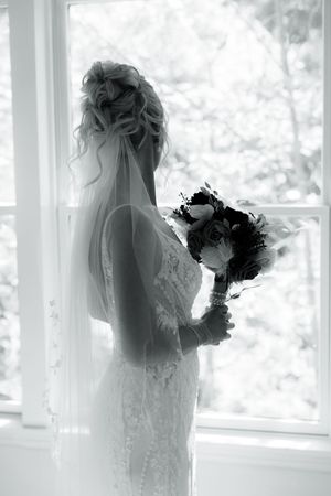 Silhouette of bride holding bouquet near bright window in wedding dress.
