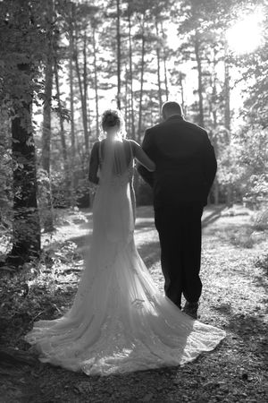 Wedding couple walking through sunlit forest path in formal attire.