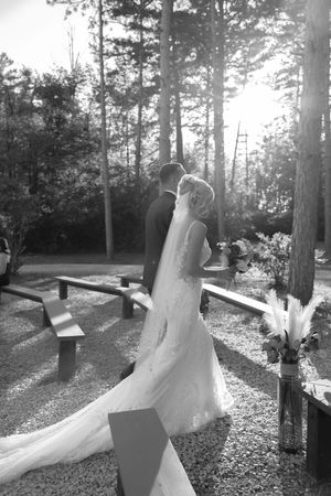 Newlyweds walking along wooded path with sunlight streaming through trees.