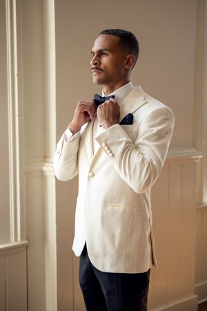Person in white tuxedo jacket adjusts bow tie in elegant indoor setting.