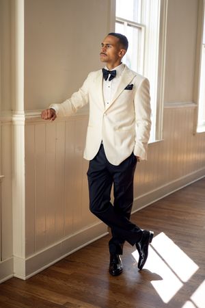 Person in formal white tuxedo poses dramatically in sunlit room with hardwood floors.