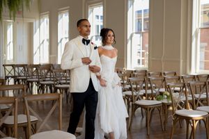 Wedding couple sharing a private moment among empty cross-back chairs in sunlit venue.
