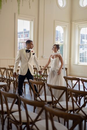 Artistic shot through wooden chairs capturing couple's first look moment in bright venue.