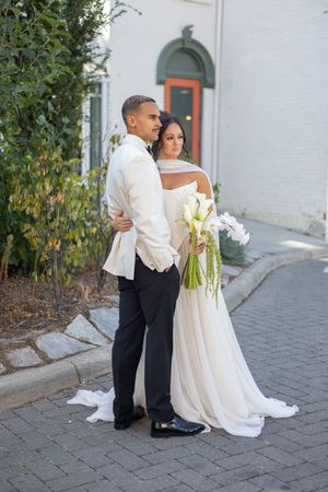 Couple embracing outdoors on brick pathway with white wedding attire and floral bouquet.