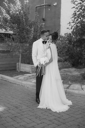 A black and white wedding photo shows a couple posing on a brick patio with greenery in the background.