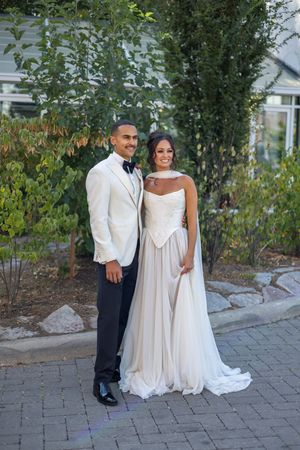 A couple in elegant white wedding attire poses together on a stone paved pathway near garden landscaping.