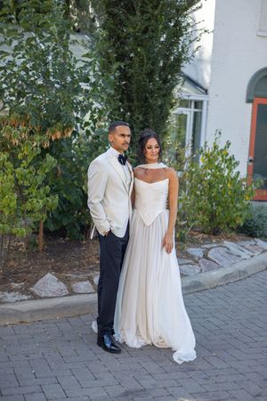 Newlyweds in white attire pose for romantic portraits on a stone walkway near landscaped gardens.