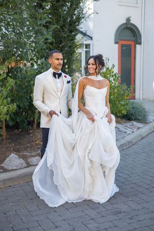 Wedding couple holds their formal white attire while posing together on brick pavers outdoors.