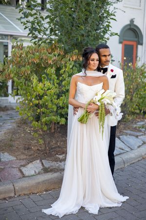 Couple in white wedding attire poses with floral bouquet on stone path near garden landscaping.
