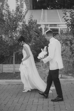 A couple in wedding attire walk along a brick path in a garden setting, captured in black and white photography.