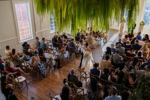 Wedding ceremony in a bright indoor venue with hanging greenery and wooden floors filled with seated guests.