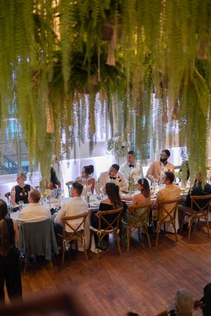 Elegant dinner party under hanging greenery with guests seated at a long table in an indoor venue.