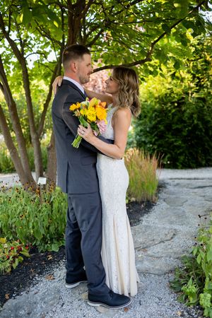 Bride and groom embrace on garden path surrounded by lush greenery.