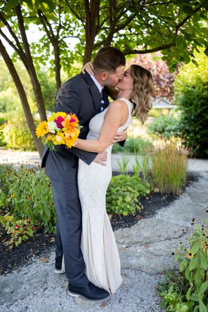 Romantic wedding couple embraces on a garden path surrounded by lush greenery and bright summer flowers.