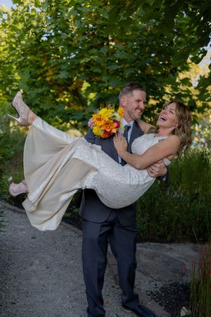 Groom lifts bride in white lace dress during playful garden photo session.