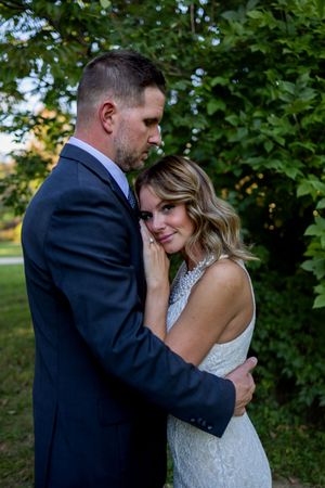 Romantic outdoor wedding portrait of couple embracing under green trees in evening light.
